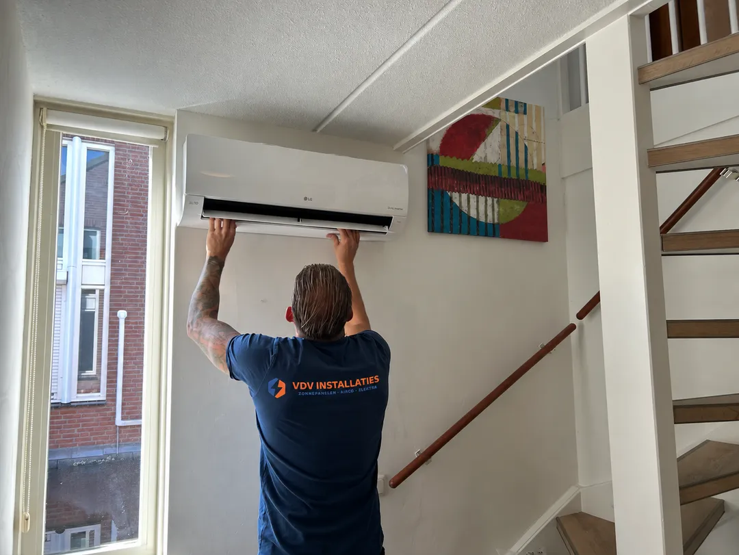 Een monteur van VDV Installaties, met het logo op zijn rug, hangt een airco binnenunit op in een trapgat
